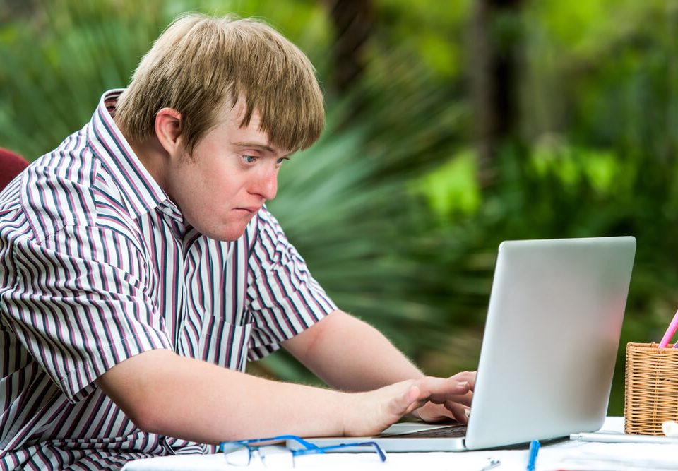 Jovem sentado com Síndrome de Down digitando num laptop. Mostrando a capacidade de superação e vontade de socializar-se.