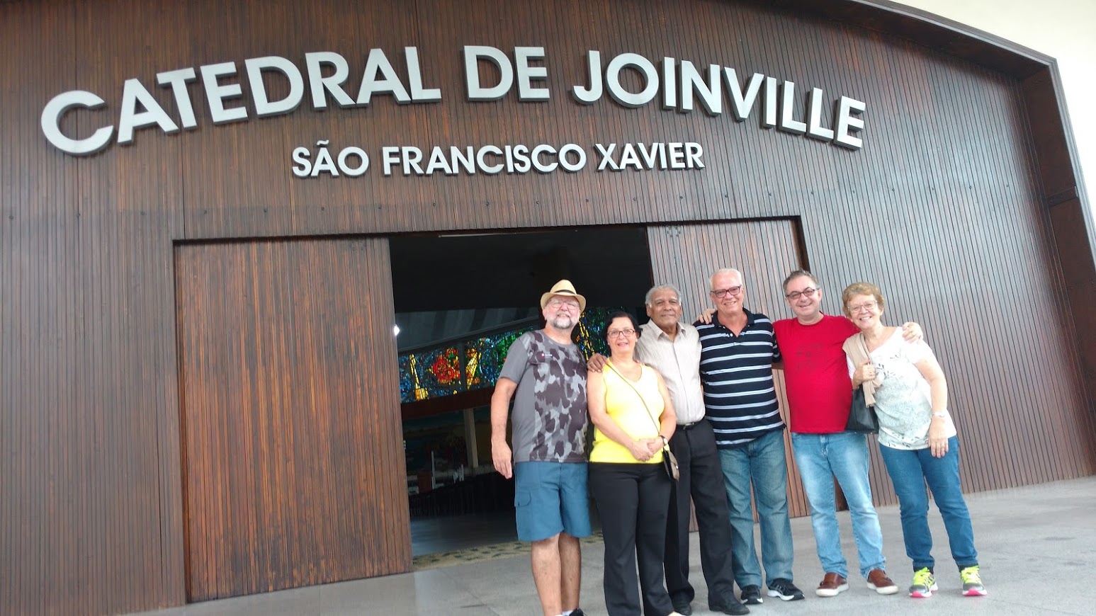 Sete surdos em frente à Catedral de Joinville. Fachada de madeira escura, e escrito Catedral de Joinville, São Francisco Xavier.