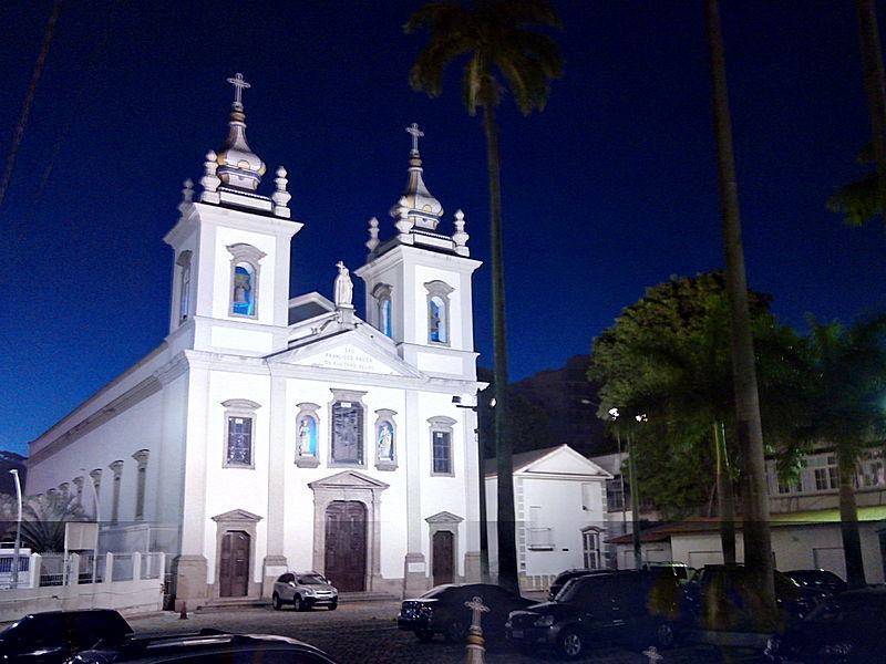 Foto da Igreja de São Francisco Xavier - Tijuca - duas grandes torres, de cor branca e nave central bem longa. Em frente com coqueiros e estacionamento em frente da Igreja e com alguns carros.